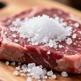 Sal grosso sendo espalhado sobre um bife cru em uma tábua de madeira, pronto para ir à churrasqueira